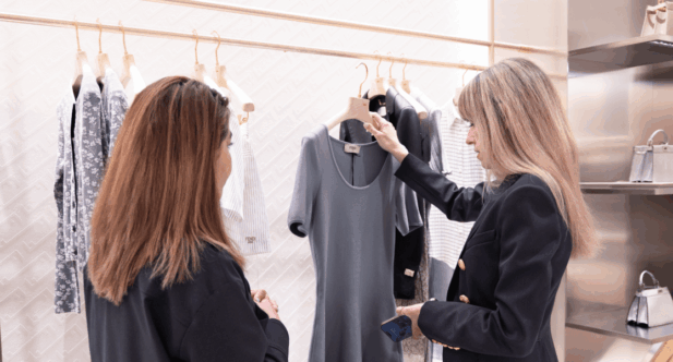 Raquel Macias, personal stylist showing a gray dress to a client inside a modern boutique, with clothing neatly displayed on wooden hangers in the background.
